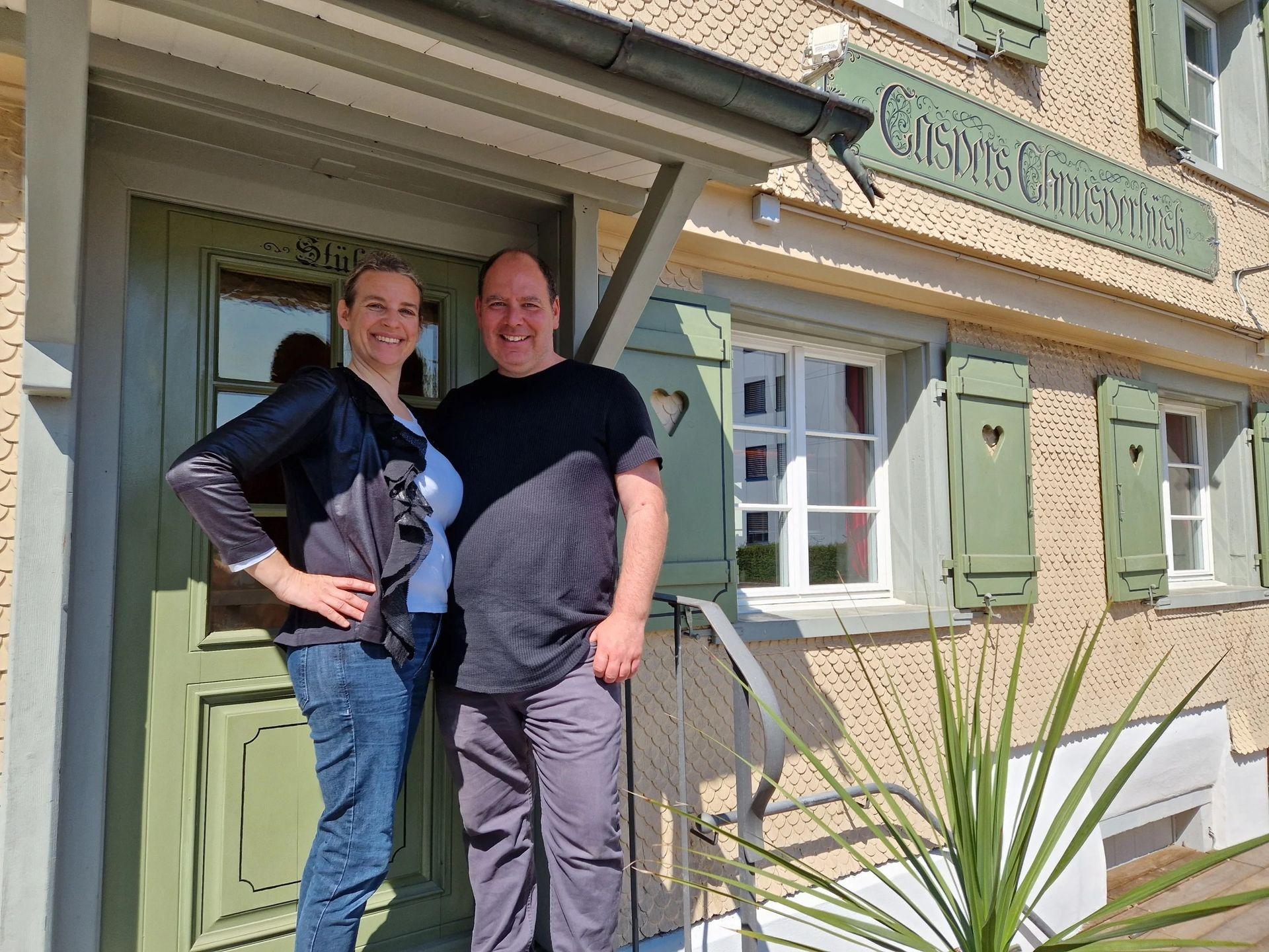 Two people smiling in front of a building with green shutters and a sign, standing on steps.